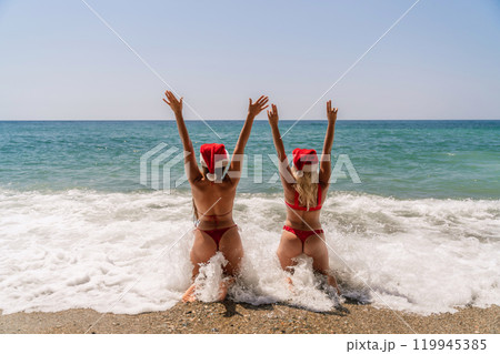 Women Santa hats ocean play. Seaside, beach daytime, enjoying beach fun. Two women in red swimsuits and Santa hats are enjoying themselves in the ocean, kneeling in the waves and raising their hands 119945385