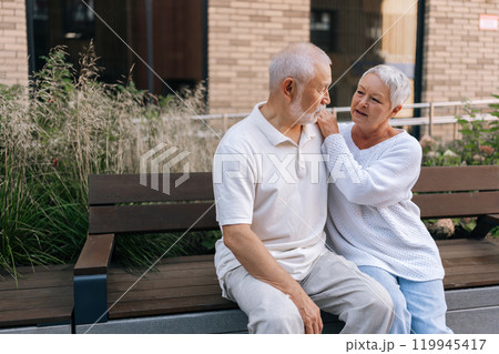 Beautiful affectionate elderly couple enjoying each other company sitting on bench on summer day, loving looking at each other, showcasing beauty of enduring love and companionship. Beautiful affectionate elderly couple enjoying each other company sitting on bench on summer day, loving looking at each other, showcasing beauty of enduring love and companionship. 119945417