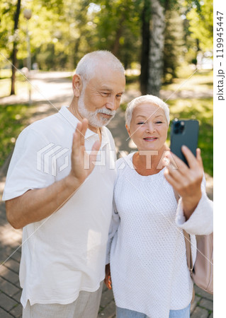 Vertical portrait of happy senior couple enjoying video call in summer sunny park, having smiling, laughing and cherishing moments in retirement, embracing technology and staying connected outdoors. Vertical portrait of happy senior couple enjoying video call in summer sunny park, having smiling, laughing and cherishing moments in retirement, embracing technology and staying connected outdoors. 119945447