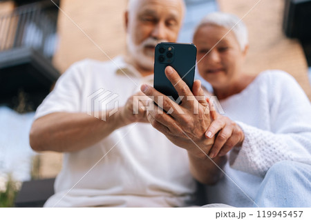 Selective focus shot of elderly married couple using smartphone, exploring apps, staying connected with world, embracing digital age in retirement. Senior male and female typing phone together outside Selective focus shot of elderly married couple using smartphone, exploring apps, staying connected with world, embracing digital age in retirement. Senior male and female typing phone together outside 119945457