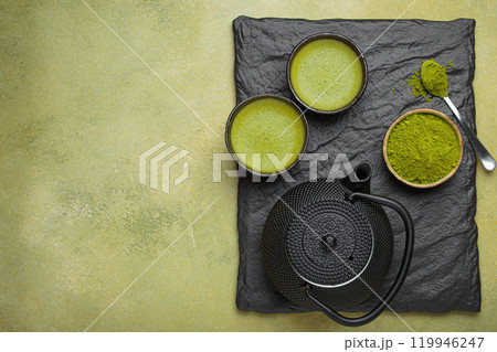 Matcha tea powder and hot drink in bowls with black iron teapot on black slate on green background, Japanese tea top view with copy space 119946247