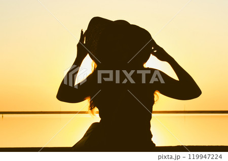 A silhouette of stylish, extravagant woman wearing a straw hat poses against a yellow sky at sunset. Travel vacation by the sea, ocean in summer nature. Female traveler back view in morning sunlight. 119947224