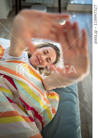 Portrait middle aged woman through palm smiling lying under blanket in bed awaked with good mood. 119947327