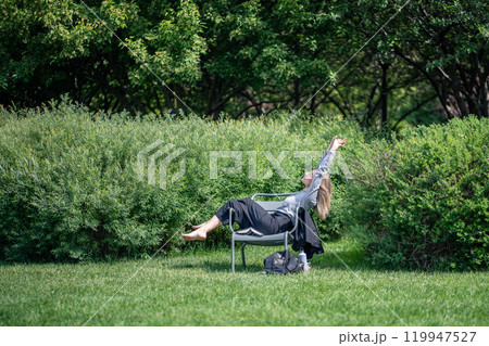 Carefree woman relaxing on park chair surround lush plants barefoot stretching arms on break outdoor Carefree woman relaxing on park chair surround lush plants barefoot stretching arms on break outdoor 119947527