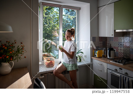 Easy living, slow morning of pleased woman in pajamas enjoying coffee while scrolling web on phone. 119947558