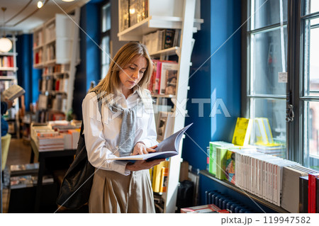 Focused thoughtful middle aged woman reading book standing shopping in modern bookstore near window Focused thoughtful middle aged woman reading book standing shopping in modern bookstore near window 119947582