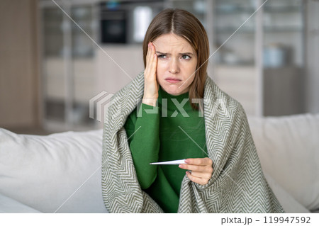 Upset sick woman with thermometer, feel headache and seasonal cold symptoms at home in autumn Upset sick woman with thermometer, feel headache and seasonal cold symptoms at home in autumn 119947592