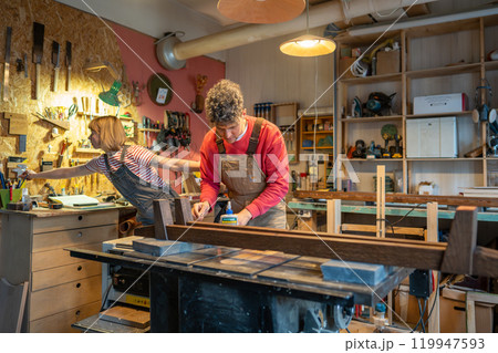 Artisan collaborating with apprentice assistant, constructs bed frame from solid wood in workshop Artisan collaborating with apprentice assistant, constructs bed frame from solid wood in workshop 119947593