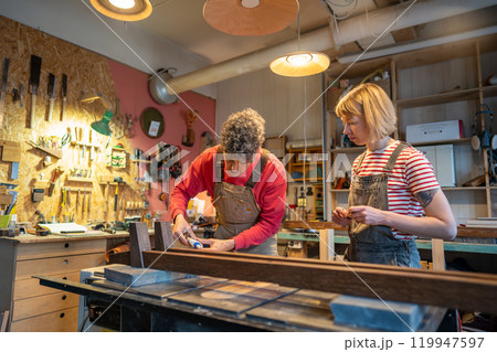 Craftsman demonstrate wooden skills to assistant in workshop. Master work on bed frame, apply paste Craftsman demonstrate wooden skills to assistant in workshop. Master work on bed frame, apply paste 119947597