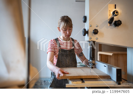 Female joiner use jigsaw and lathe, cut wood piece into required shape for production in workshop 119947605