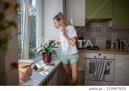 Pleased serene woman spending slowly relaxed morning time alone drinking coffee caring houseplants. 119947629