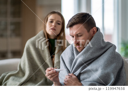Sick shivering couple of man and woman wrapped in blanket. Male measure fever on couch, battling flu Sick shivering couple of man and woman wrapped in blanket. Male measure fever on couch, battling flu 119947638
