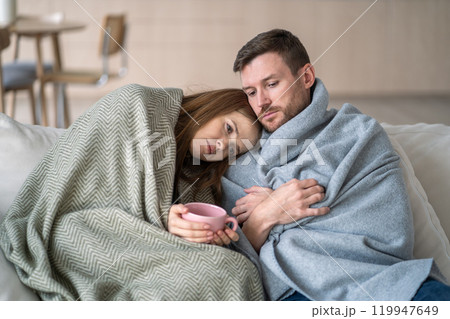 Sad tired couple wrapped in blanket on couch, drink hot tea at home, feeling sick during cold season Sad tired couple wrapped in blanket on couch, drink hot tea at home, feeling sick during cold season 119947649
