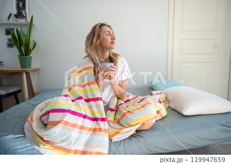 Relaxed middle aged woman wrapped in blanket, holding mug of morning coffee in bed looks at window. 119947659