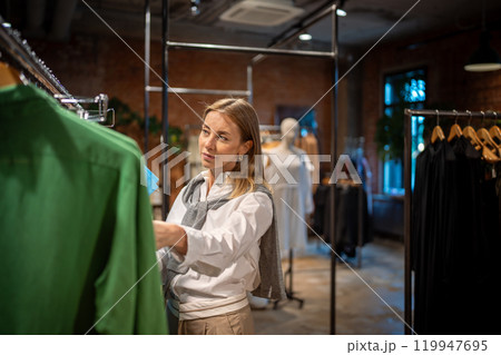 Pleased woman shopaholic examines clothing with interest before purchase, making choice in boutique. Pleased woman shopaholic examines clothing with interest before purchase, making choice in boutique. 119947695