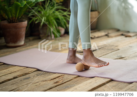 African woman stands on gym mat using massage ball to roll out tension feet spend time for self-care 119947706