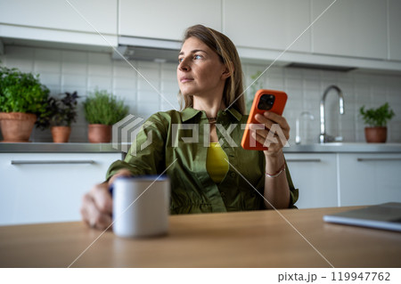 Thoughtful woman sits in kitchen with cup holding smartphone immersed daydreaming looking to window. Thoughtful woman sits in kitchen with cup holding smartphone immersed daydreaming looking to window. 119947762