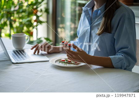 Peaceful woman at table in morning holding sandwich having breakfast using laptop computer. 119947871