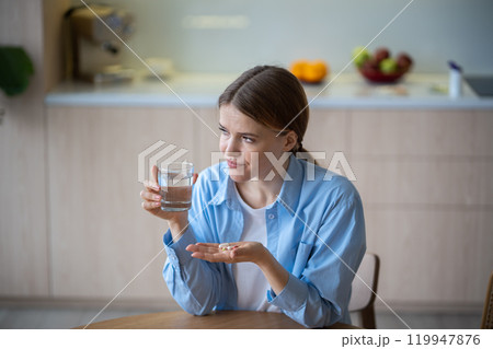 Stressed unhealthy woman holding capsules, glass of water, suffering from chronic pain, disease 119947876