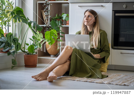 Pensive woman with laptop looking at window, feeling lack of energy sit on floor at home with plants Pensive woman with laptop looking at window, feeling lack of energy sit on floor at home with plants 119947887