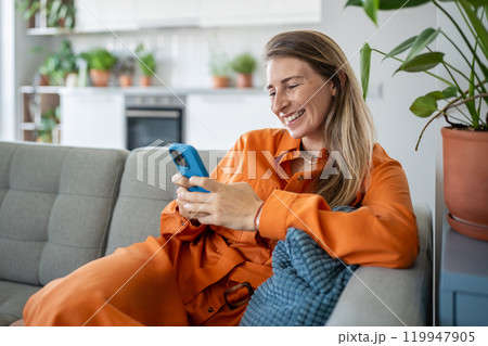 Happy Swedish woman using smartphone, resting at sofa at cozy home and scrolling social networks Happy Swedish woman using smartphone, resting at sofa at cozy home and scrolling social networks 119947905