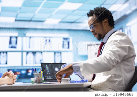 African american medic preparing for patient examination during check up visit, putting on his disposable rubber gloves in cabinet. Doctor reviewing diagnostic reports and results. 119949669