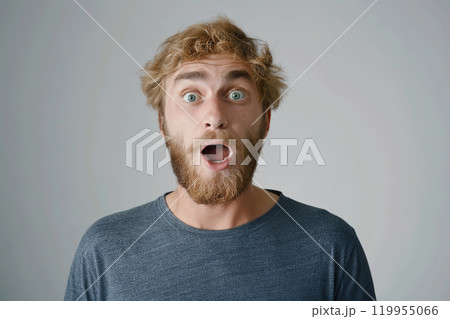 A very excited, surprised man looks at the camera with his mouth open, isolated on a light gray background. A funny-looking red-haired and bearded man. Live emotions. Idea for advertising. Copy space 119955066