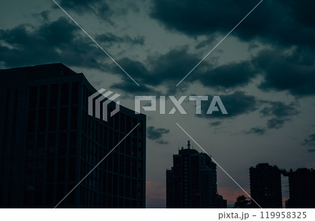 Silhouettes of tall multi-story buildings against a dark blue cloudy sky at sunset dusk. View of tall residential houses in a big city in a twilight. 119958325