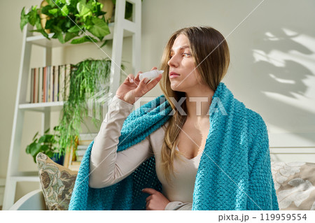 Young woman with runny nose using nasal spray sitting on couch at home. 119959554