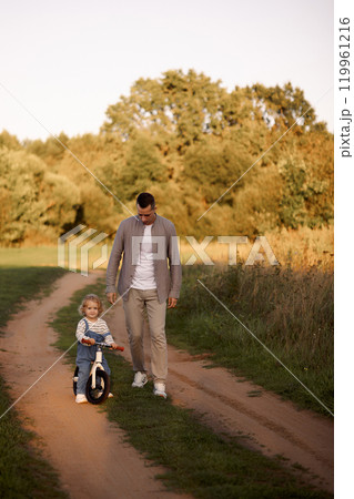 Father teaches a young child to ride a balance bike in the park in the summer Father teaches a young child to ride a balance bike in the park in the summer 119961216