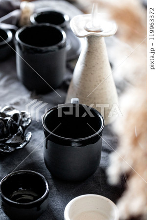 Rustic table setting with empty craft handmade ceramic tableware, black and white rough bowls, cups, jug and vase on linen tablecloth. Dry reed flowers. Day light dining room. 119963372