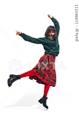 Energetic woman in green sweater and plaid skirt jumps with arms raised, embodying joy and freedom in urban fashion against white studio background. Energetic woman in green sweater and plaid skirt jumps with arms raised, embodying joy and freedom in urban fashion against white studio background. 119965511