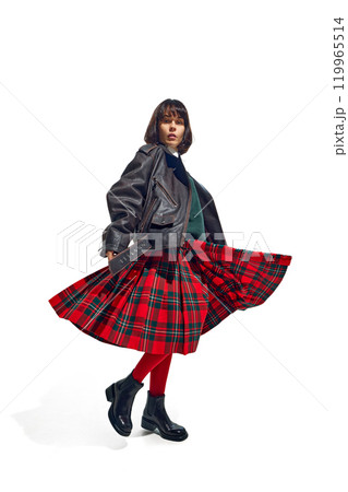 Vibrant and stylish young pretty woman in plaid skirt and leather jacket caught mid-twirl against white studio background. 119965514