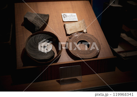 Antique film reels and Kinora projector discs displayed in a dimly lit storage box at a historical exhibit in London. Old film reels and Kinora projector discs are arranged neatly in a dark wooden box 119968341