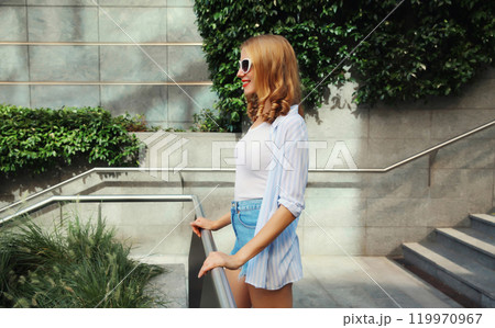 Beautiful modern young woman looking away standing on city street in summer wearing shorts, shirt Beautiful modern young woman looking away standing on city street in summer wearing shorts, shirt 119970967
