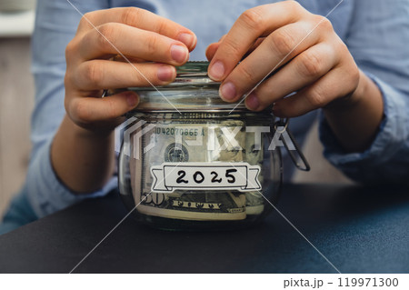 Female hands holding Glass jar full of American currency dollars cash banknote with text 2025 year. Preparation saving money. Moderate consumption and economy Female hands holding Glass jar full of American currency dollars cash banknote with text 2025 year. Preparation saving money. Moderate consumption and economy 119971300