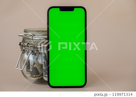 Vertical Green screen on modern mobile phone in background of glass jar full of American currency dollar banknotes on beige background. Cope space for text. Advertisement for application website 119971314