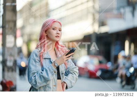 A woman with pink hair is standing on a street holding a cell phone 119972612