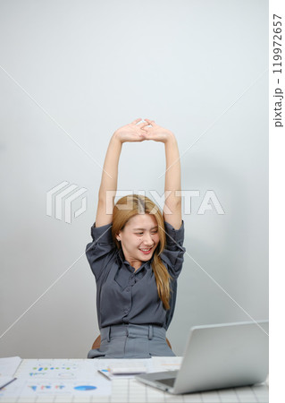 A woman is stretching her arms and smiling while sitting at a desk with a laptop 119972657