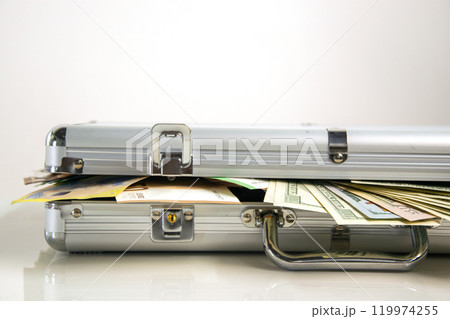 A close-up of an open silver aluminum case with cash spilling out from inside. The case is on a white surface with the cash slightly overlapping the edge 119974255