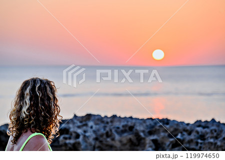 Blonde curly hair woman looking at a sunset with orange sky on the beach of Menorca, Balearic Islands. Sunset, nature Blonde curly hair woman looking at a sunset with orange sky on the beach of Menorca, Balearic Islands. Sunset, nature 119974650