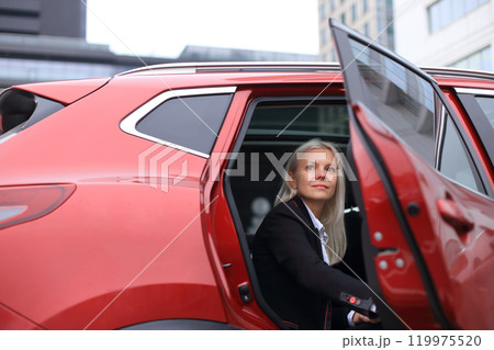 A professional businesswoman sits confidently in her stylish red car, parked outside a modern business center.  119975520