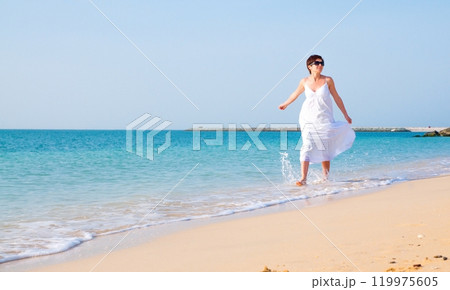 Young brunette woman in summer white dress walking on coast. Caucasian girl relaxing and enjoying peace on vacation Young brunette woman in summer white dress walking on coast. Caucasian girl relaxing and enjoying peace on vacation 119975605