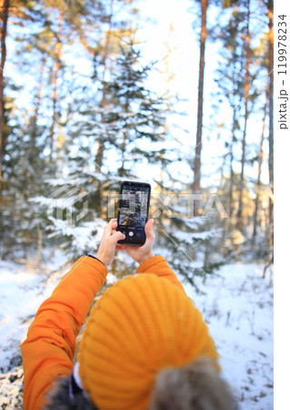 A young man stands in a winter forest, capturing the beauty of the snowy landscape with his smartphone. 119978234