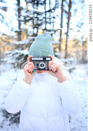 a woman holding a vintage camera, set against the stunning backdrop of a snowy winter forest. Dressed warmly for the cold, she embodies a sense of nostalgia and creativity, ready to capture the beauty 119978235