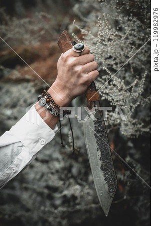 Authentic handmade knife in female hand with bracelets on atmospheric forest background in late autumn, vertical format, selective focus. 119982976