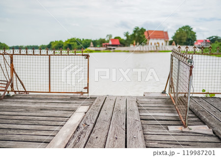 The old wooden dock with decayed iron fences beside a river in Thailand. A temple at the serene riverside. The old wooden dock with decayed iron fences beside a river in Thailand. A temple at the serene riverside. 119987201