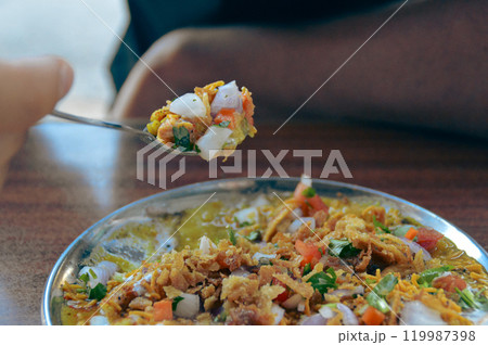 A foodie taking a spoonful of Bhel puri chaat served in a plate made of puffed rice, sev, chopped onions, and tomatoes in a vibrant mix. Traditional authentic snack food originating in Mumbai, India A foodie taking a spoonful of Bhel puri chaat served in a plate made of puffed rice, sev, chopped onions, and tomatoes in a vibrant mix. Traditional authentic snack food originating in Mumbai, India 119987398