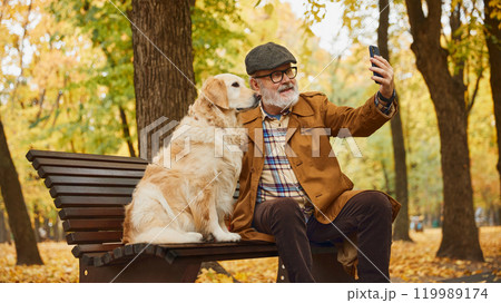 Happy mature man sitting on bench and taking selfie with his beloved pet, olden retriever dog in autumn park. 119989174