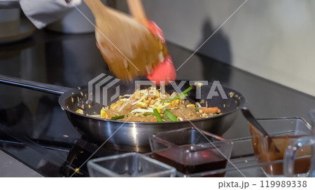 A cook stirs fry Pad Thai noodles in a pan with spatulas on electric stoves. Sauce dishes for cooking traditional Pad Thai. A cook stirs fry Pad Thai noodles in a pan with spatulas on electric stoves. Sauce dishes for cooking traditional Pad Thai. 119989338